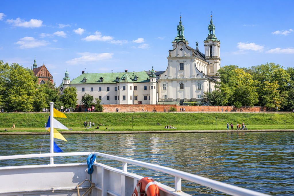 Riverside view of the Church on the Rock in Krakow from a boat on the Vistula River, with spring greenery and people walking along the embankment.