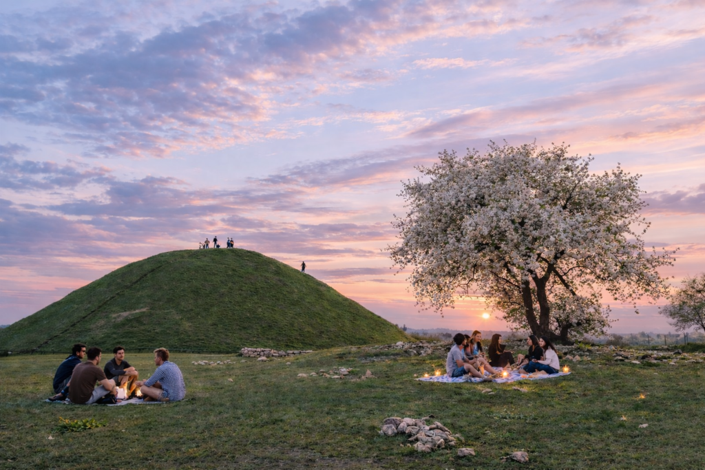 Krakus Mound in Krakow at sunset with a blooming tree and small groups of people relaxing on the grass.