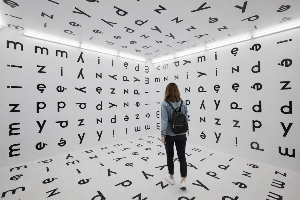 Interactive exhibition room in MOCAK Krakow with letters covering the walls, floor and ceiling, and a visitor exploring the space.
