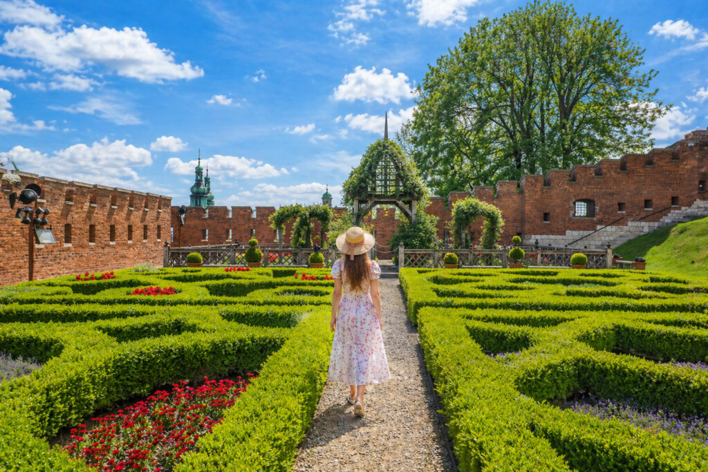 Woman in a white floral dress and summer hat walking through the formal gardens of Wawel Castle in Krakow under a bright blue sky.