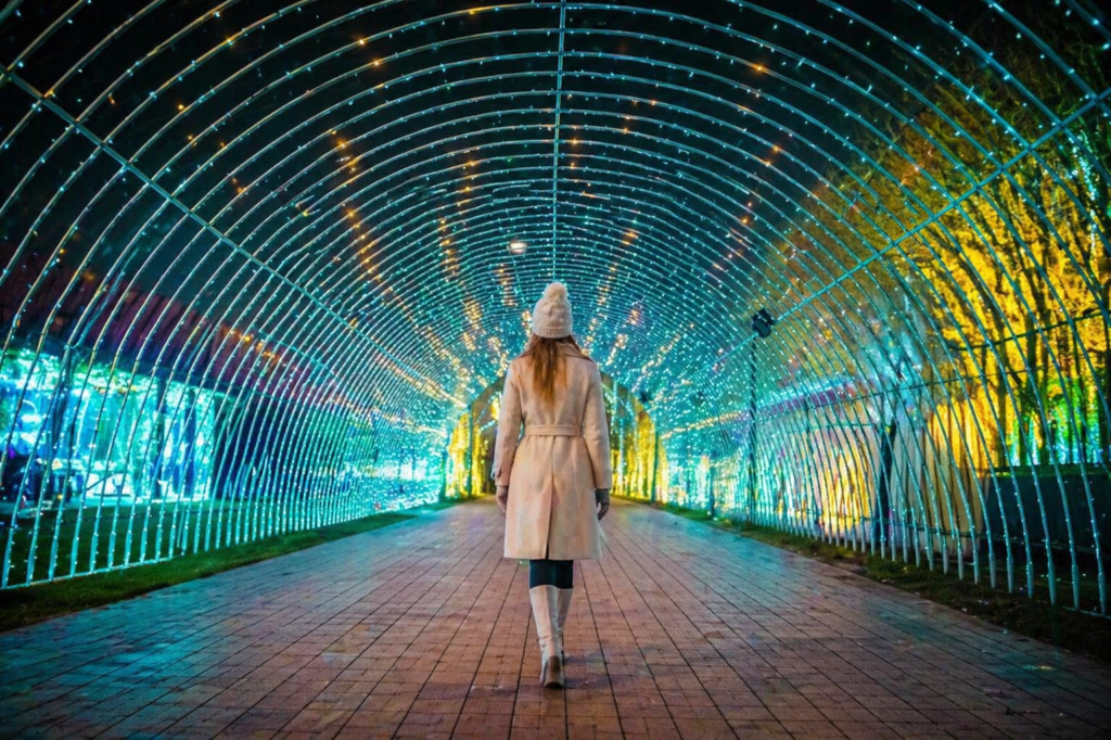 Elegant woman in a winter coat walking through a glowing light tunnel at the Garden of Lights in Krakow, viewed from behind in a symmetrical perspective.