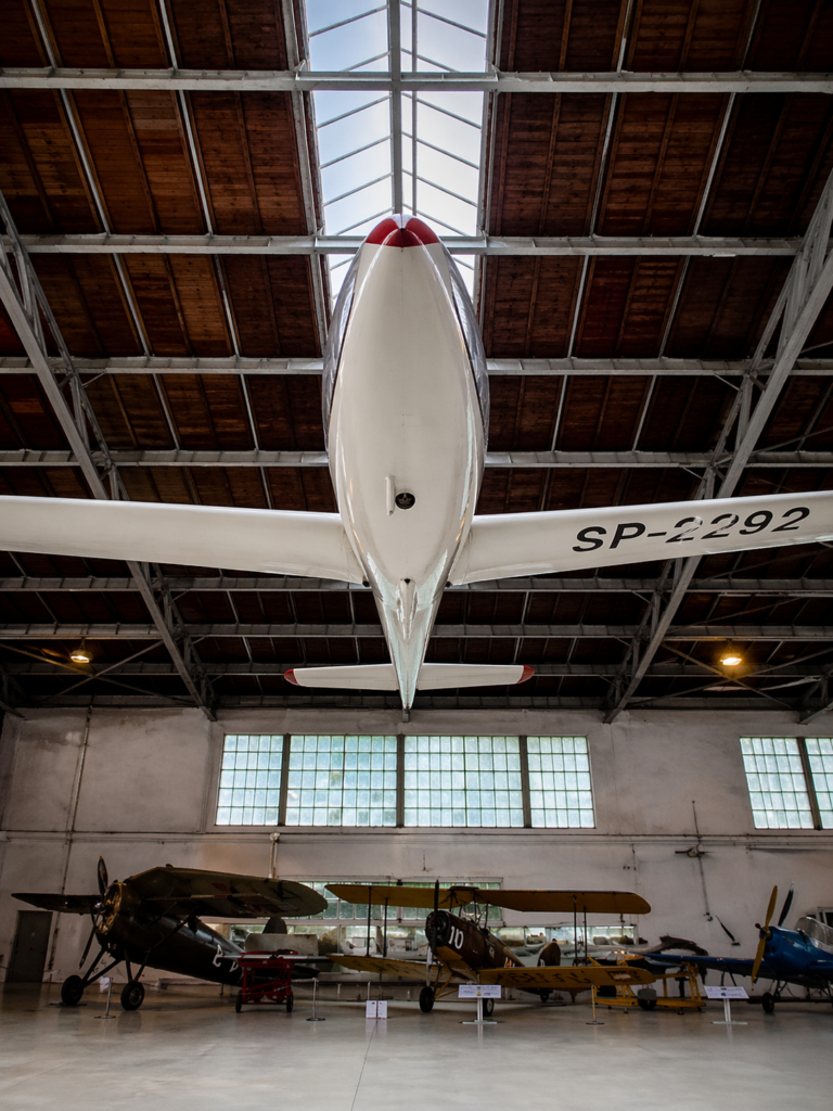 Glider aircraft in the Polish Aviation Museum in Krakow, viewed from below with a front-facing perspective highlighting the suspended plane and hangar ceiling