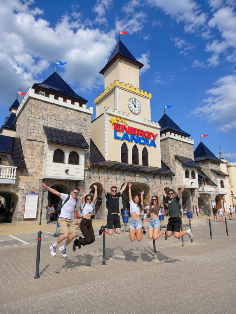 Group of friends jumping and posing for a photo in front of the main entrance to Energylandia amusement park in Poland.